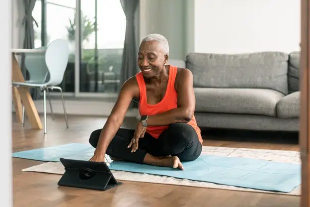 Senior woman seated on a yoga mat in a living room, looking at a tablet computer